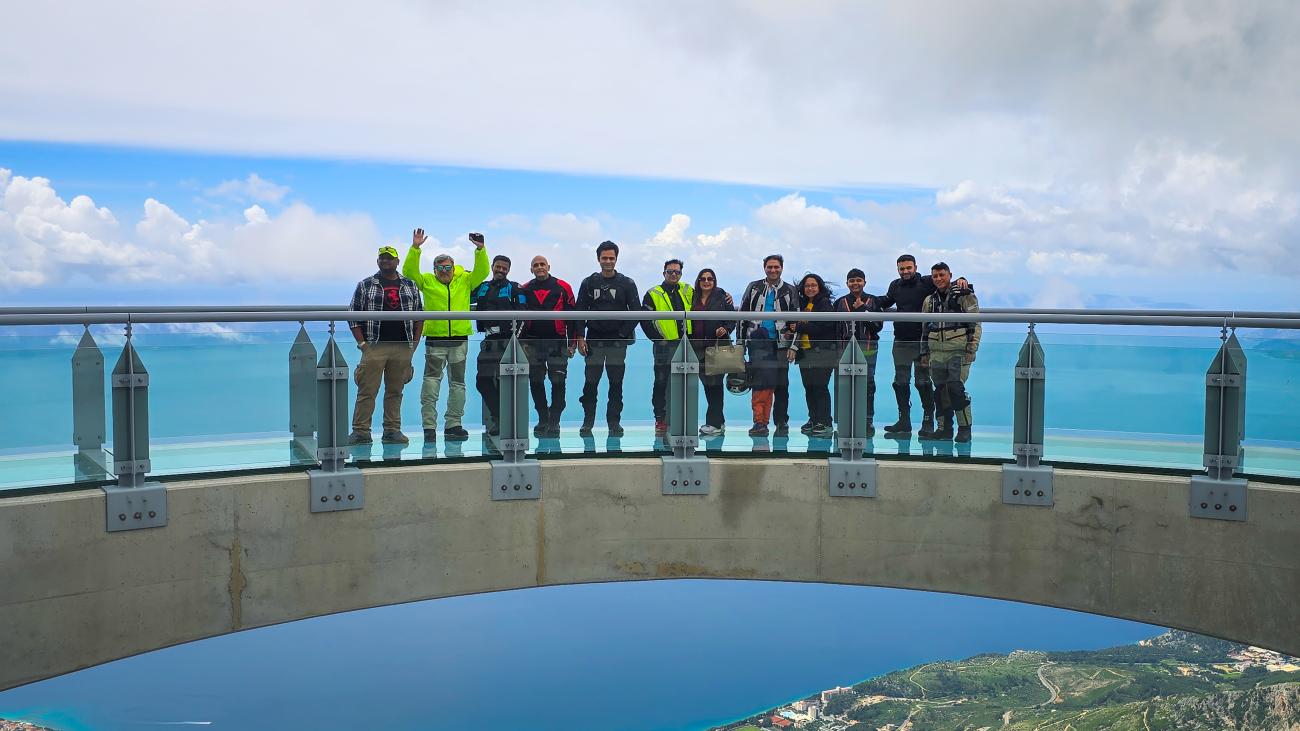 Tour group posing on a glass skywalk viewpoint high above the turquoise Balkan coastline.