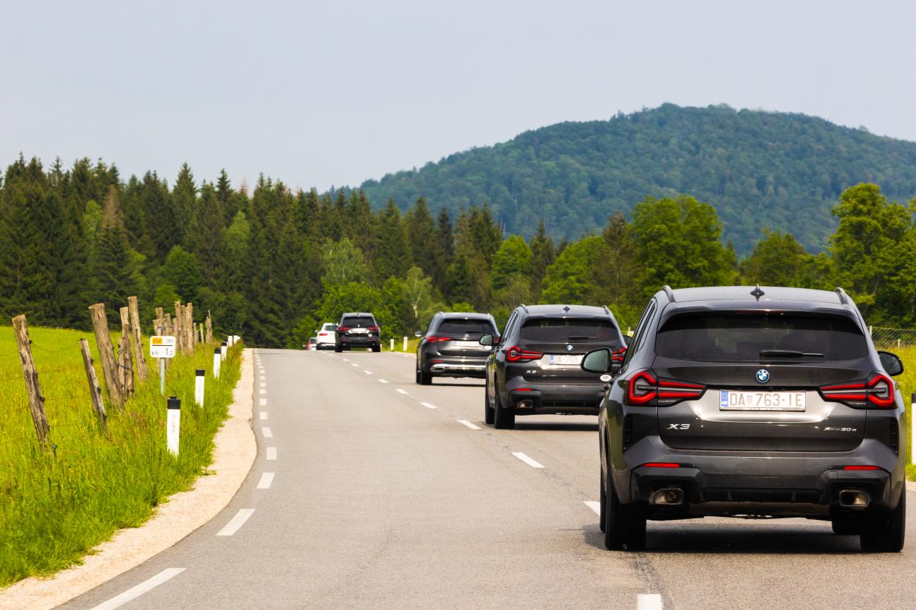 Convoy of BMW X3 SUVs driving through lush green Balkan countryside with forested hills