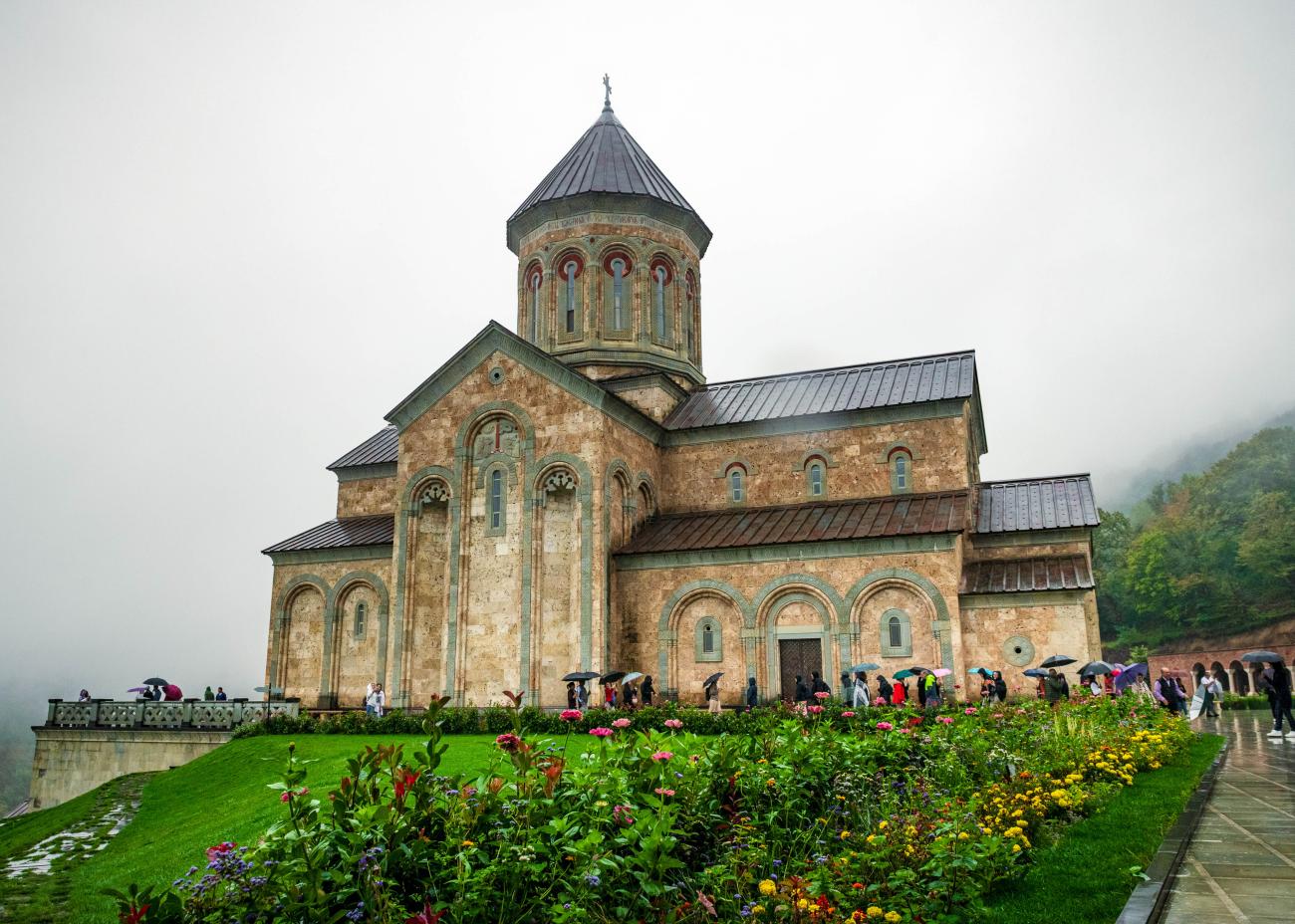 Historic Georgian monastery surrounded by flower gardens and misty Caucasus mountains.