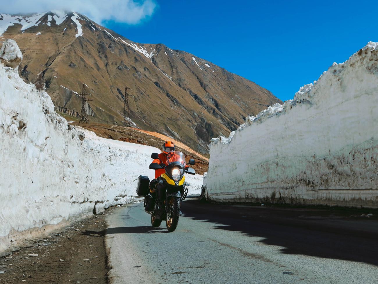 Motorcyclist in orange gear riding between towering snow walls on a high Caucasus mountain pass.