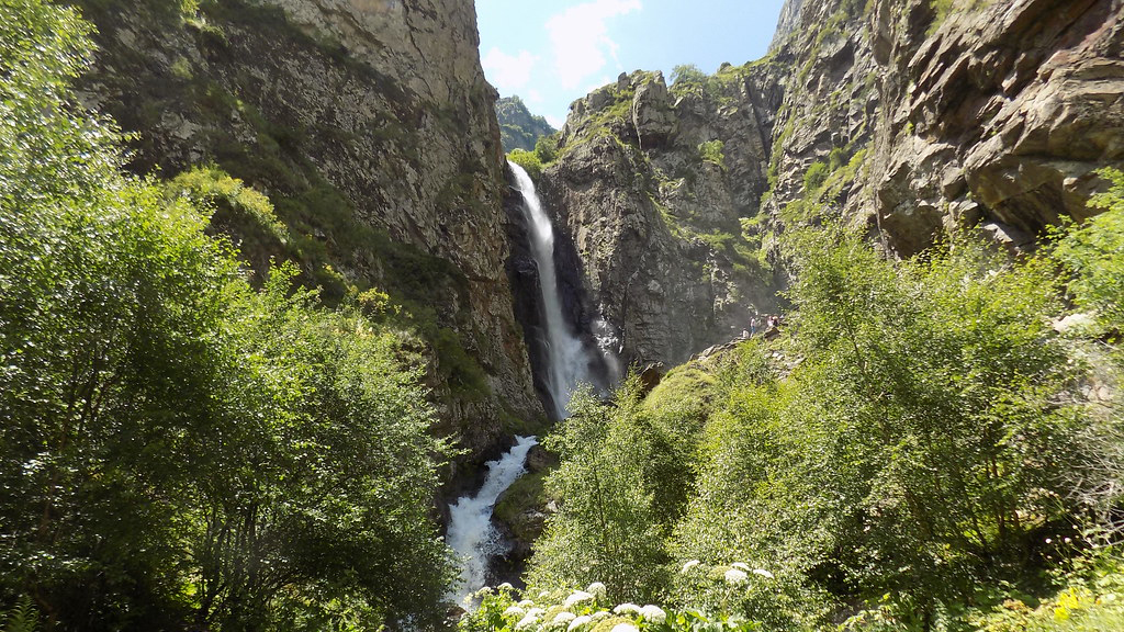 A tall waterfall cascading down a narrow rocky gorge surrounded by lush green vegetation in the Georgian Caucasus mountains