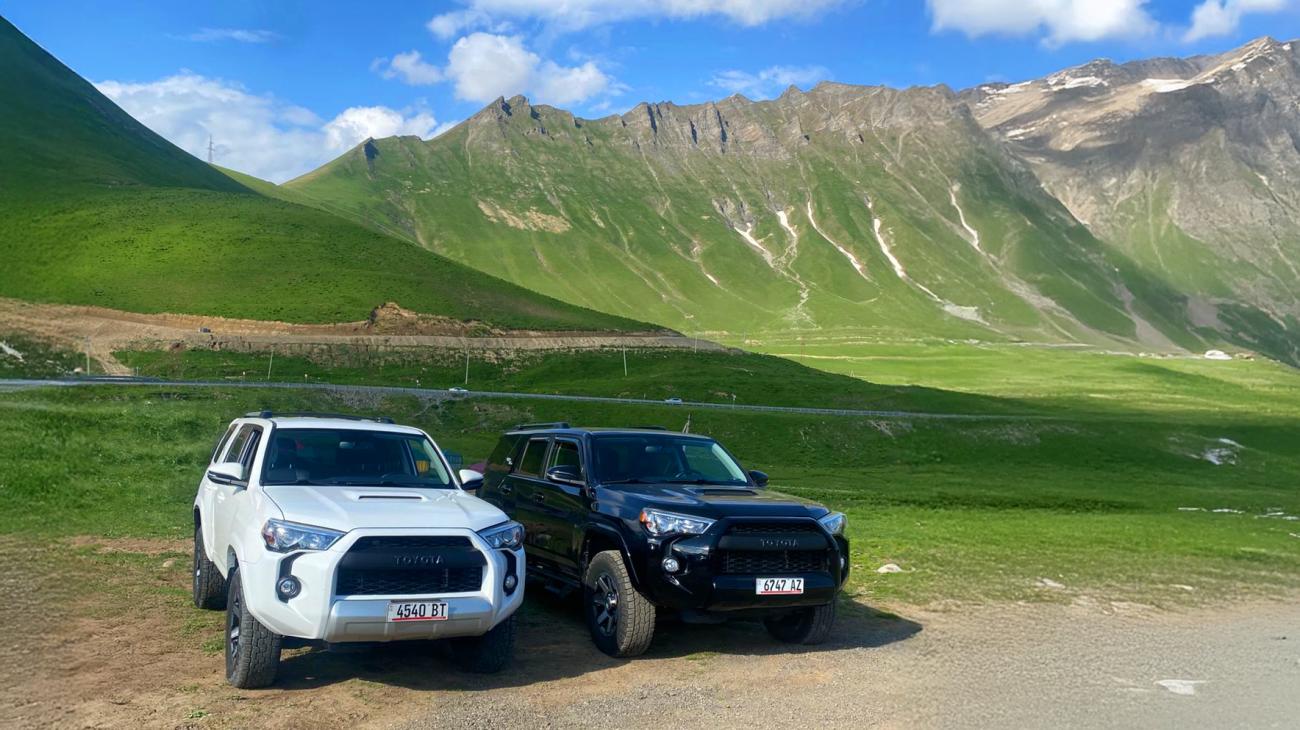 Two Toyota 4Runners parked on a mountain pass road with sweeping green valleys and rugged Caucasus ridges in the background