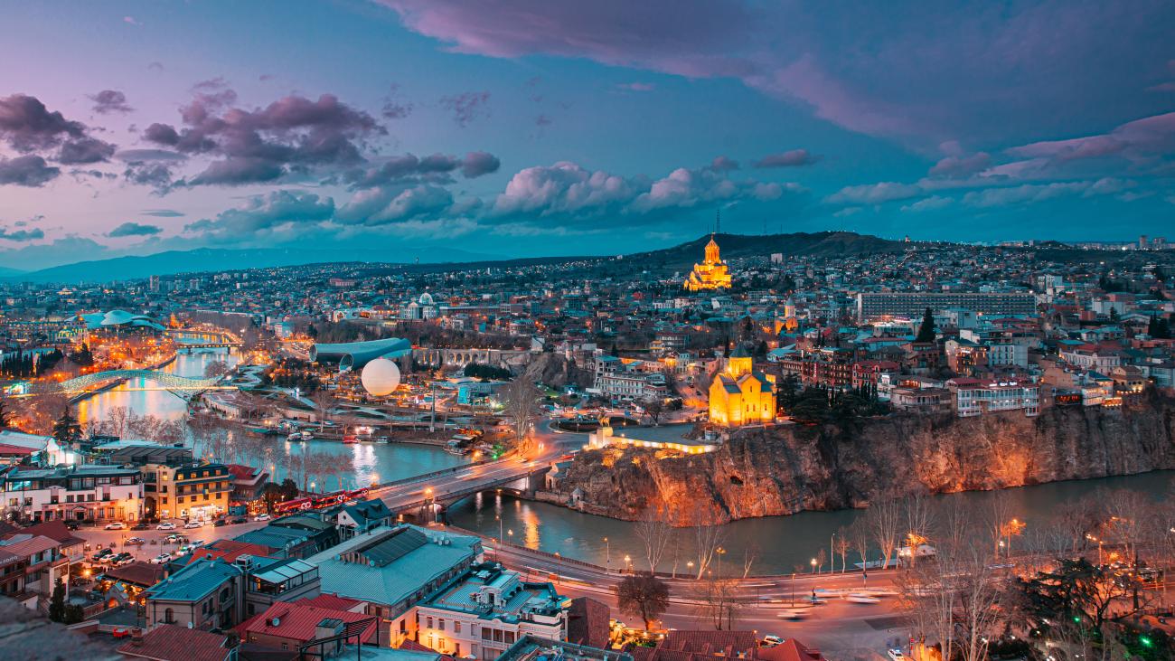 Aerial view of Tbilisi at twilight with the illuminated Holy Trinity Cathedral, Kura River, and Peace Bridge under a dramatic purple sky