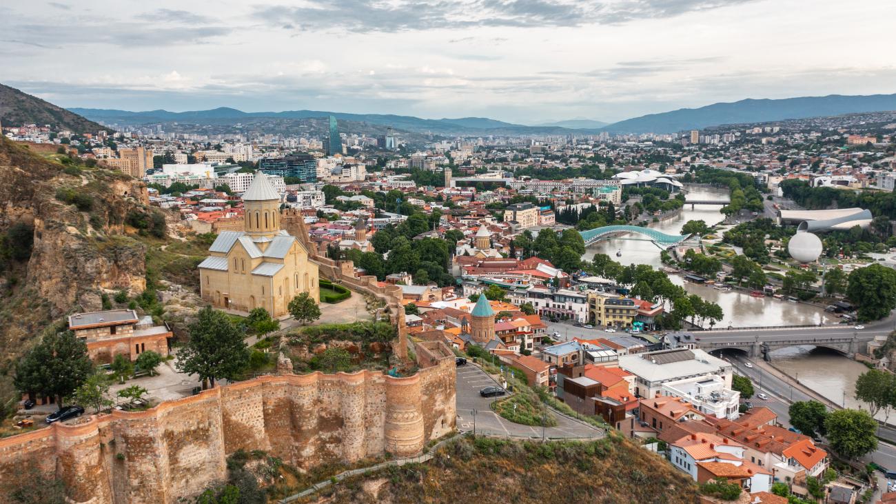 Panoramic view of Tbilisi from above showing Metekhi Church, the Peace Bridge, and the Kura River winding through the city