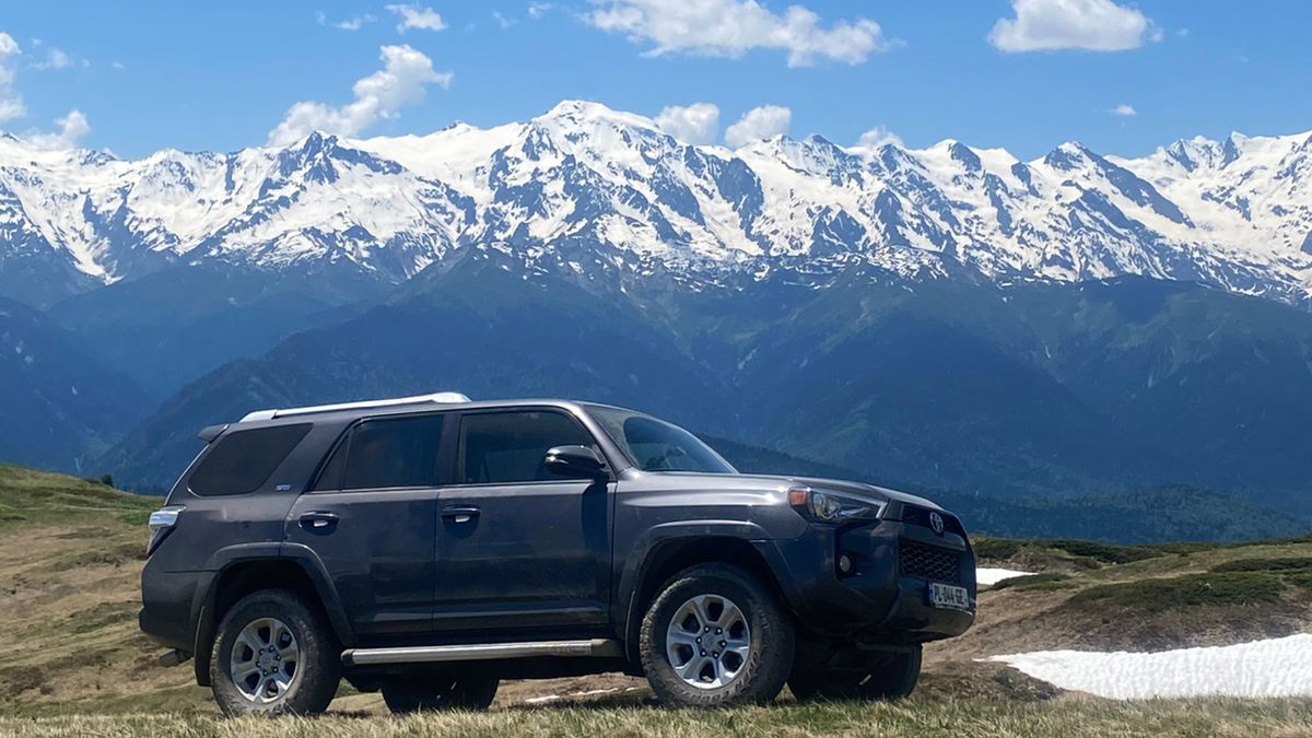 A grey Toyota 4Runner parked on a grassy alpine ridge overlooking the snow-capped Greater Caucasus mountain range in Georgia