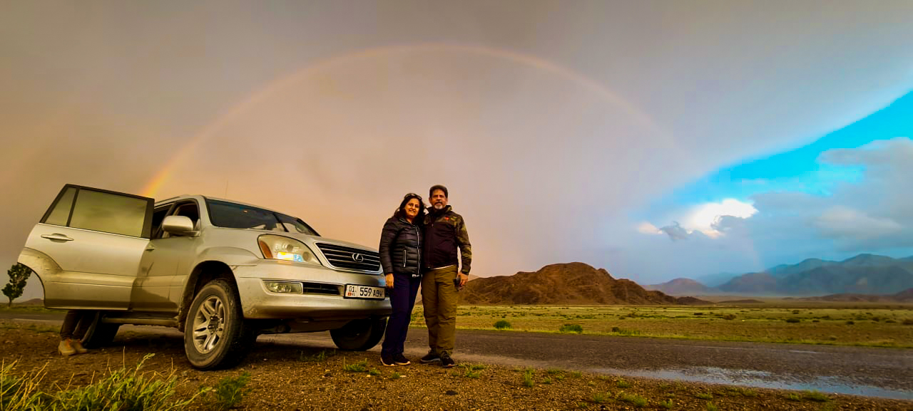 Couple posing with SUV under rainbow on Kyrgyzstan steppe