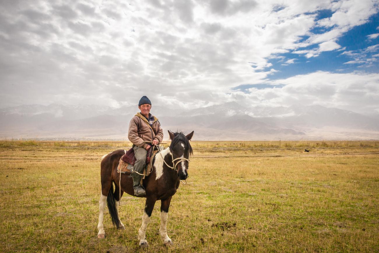 A Kyrgyz horseman sits on his horse in a wide golden steppe with misty mountains stretching across the horizon.