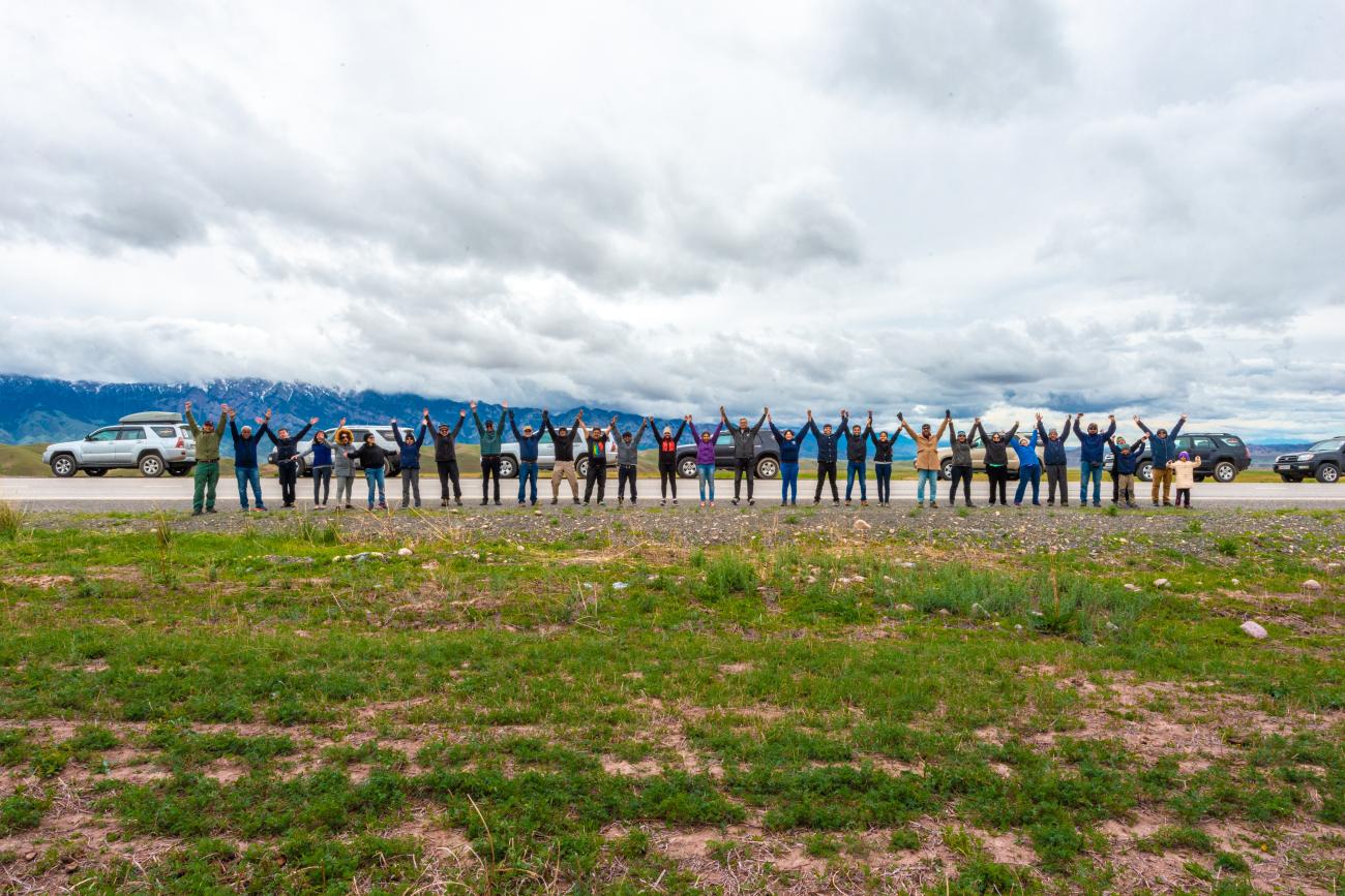 Tour group stands in a long line with arms raised on a mountain road with SUVs parked on either side.