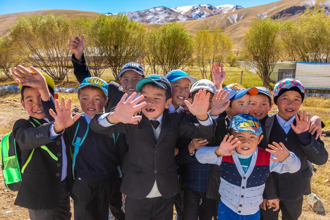 A group of smiling Kyrgyz schoolchildren wave enthusiastically at the camera in a mountain village.