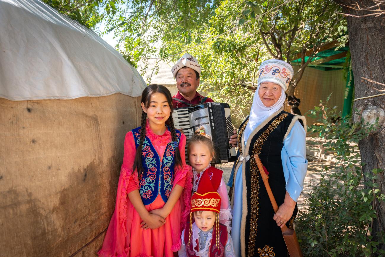 A Kyrgyz family wearing colorful traditional costumes poses beside a yurt with an accordion player.