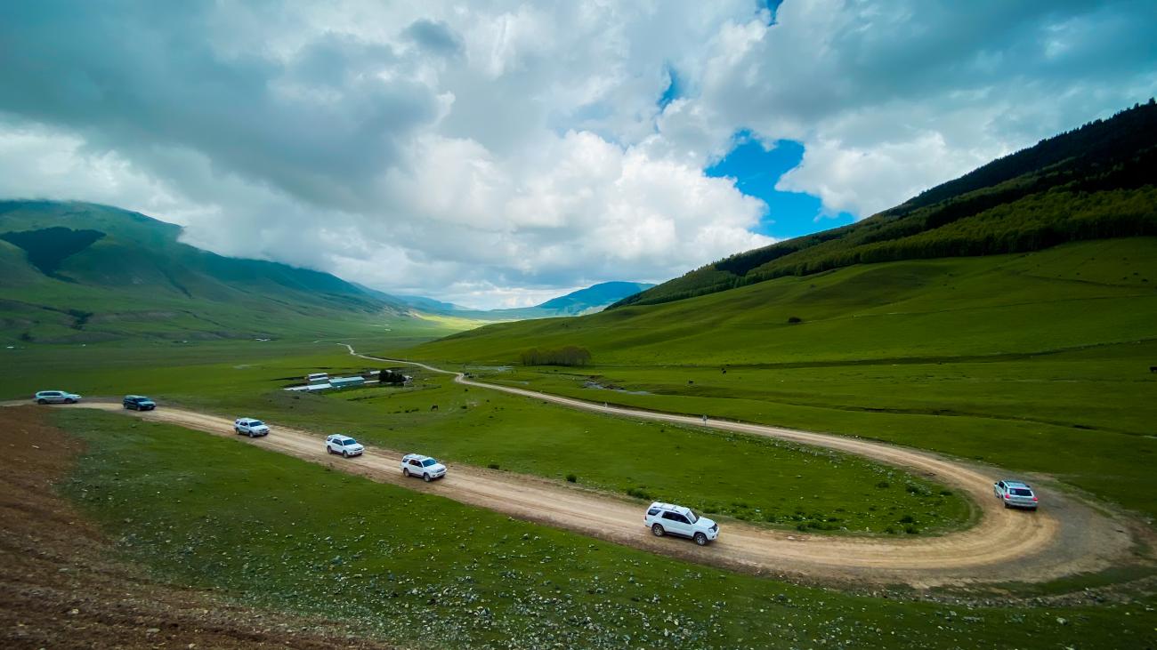 Aerial view of SUVs winding along a dirt road through a broad green valley flanked by rolling Kyrgyz mountains.