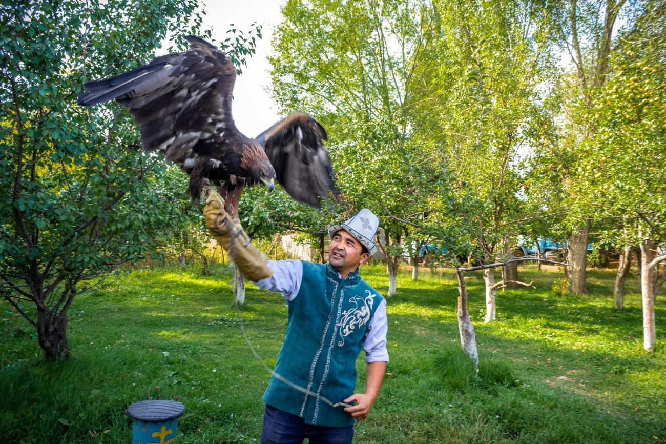 A Kyrgyz man in traditional attire holds a large eagle on his arm during a falconry demonstration in a green garden.