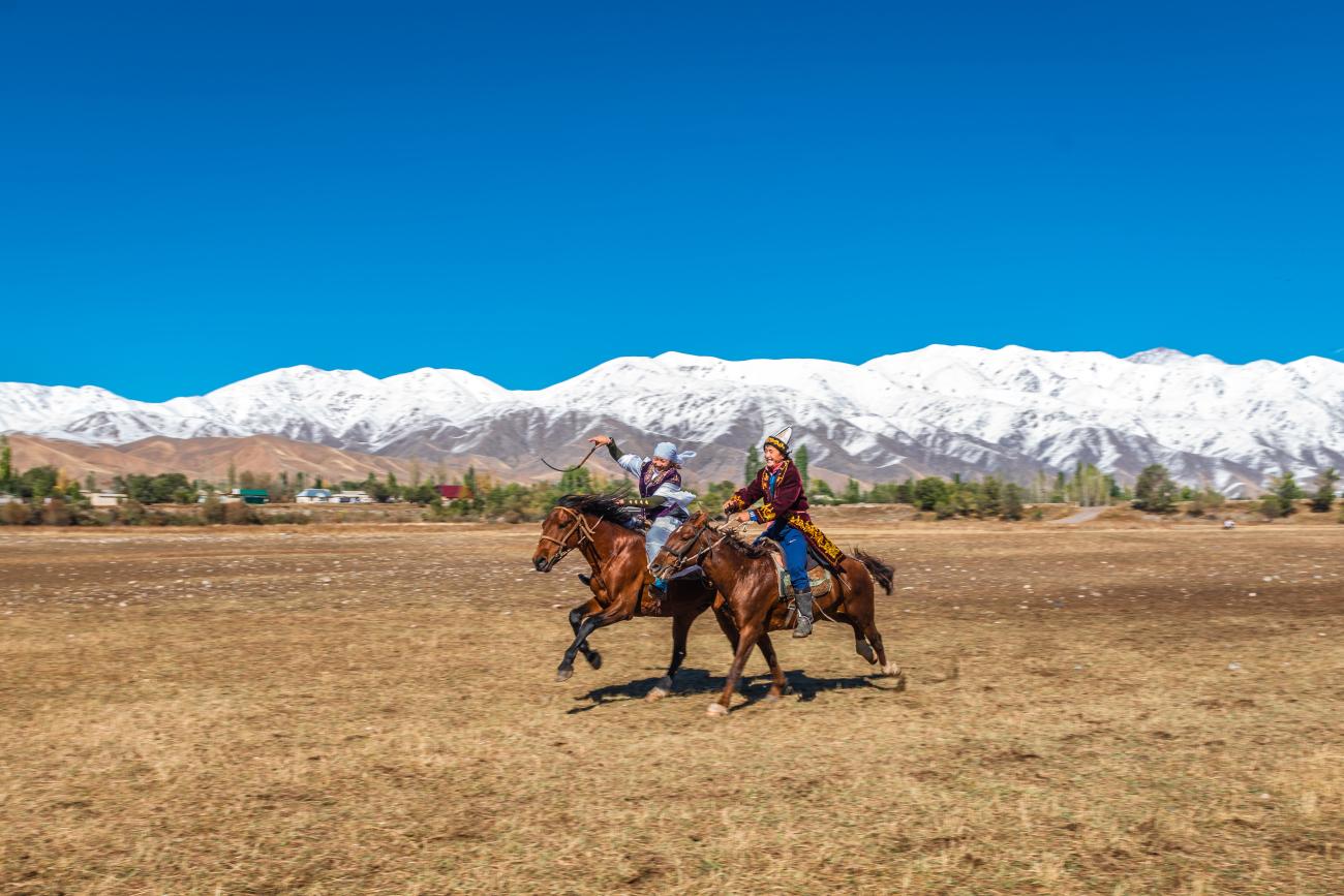 Two Kyrgyz riders in traditional dress galloping on horseback across an open plain with snow-covered mountains in the distance.