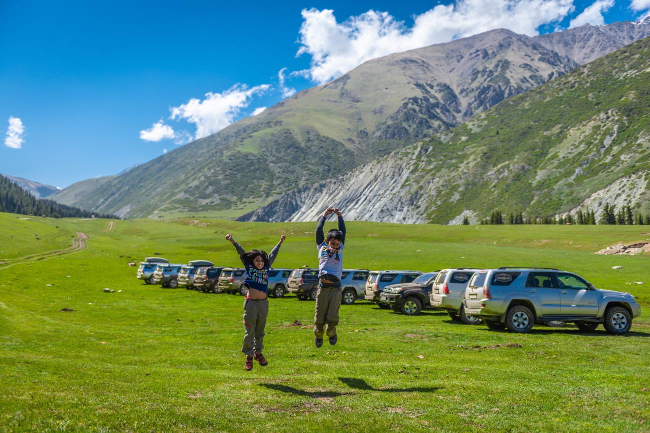 Two travelers jump for joy on a green mountain meadow in Kyrgyzstan with parked SUVs and towering peaks behind them.