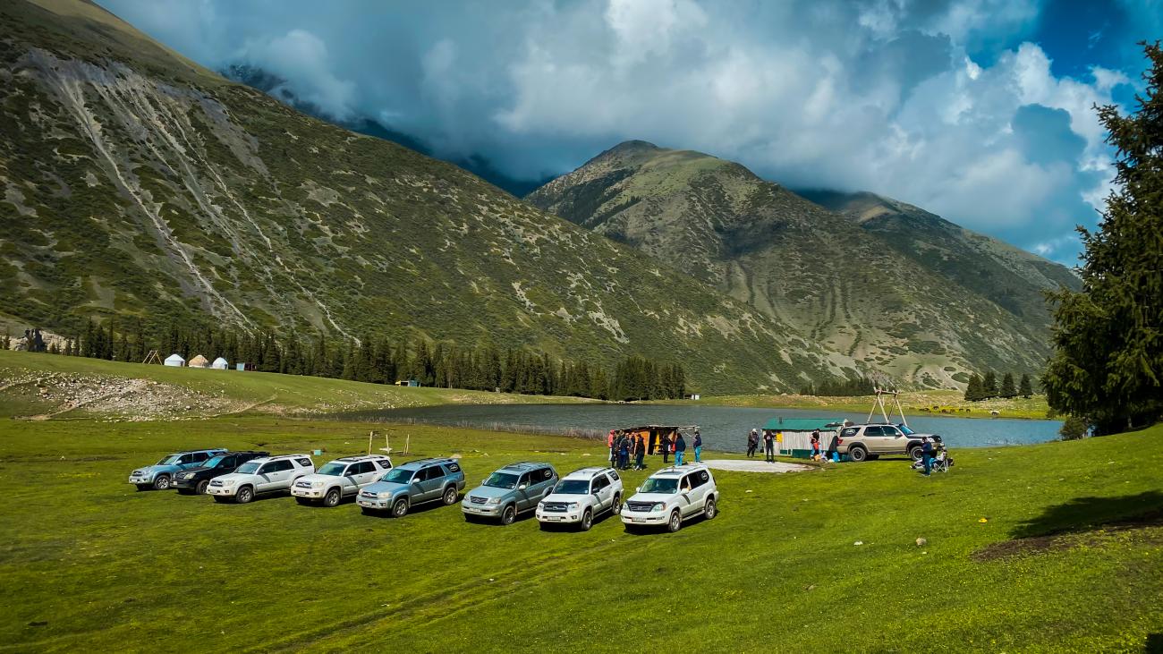 A row of SUVs parked on green grass beside an alpine lake surrounded by tall mountains and yurts in Kyrgyzstan.