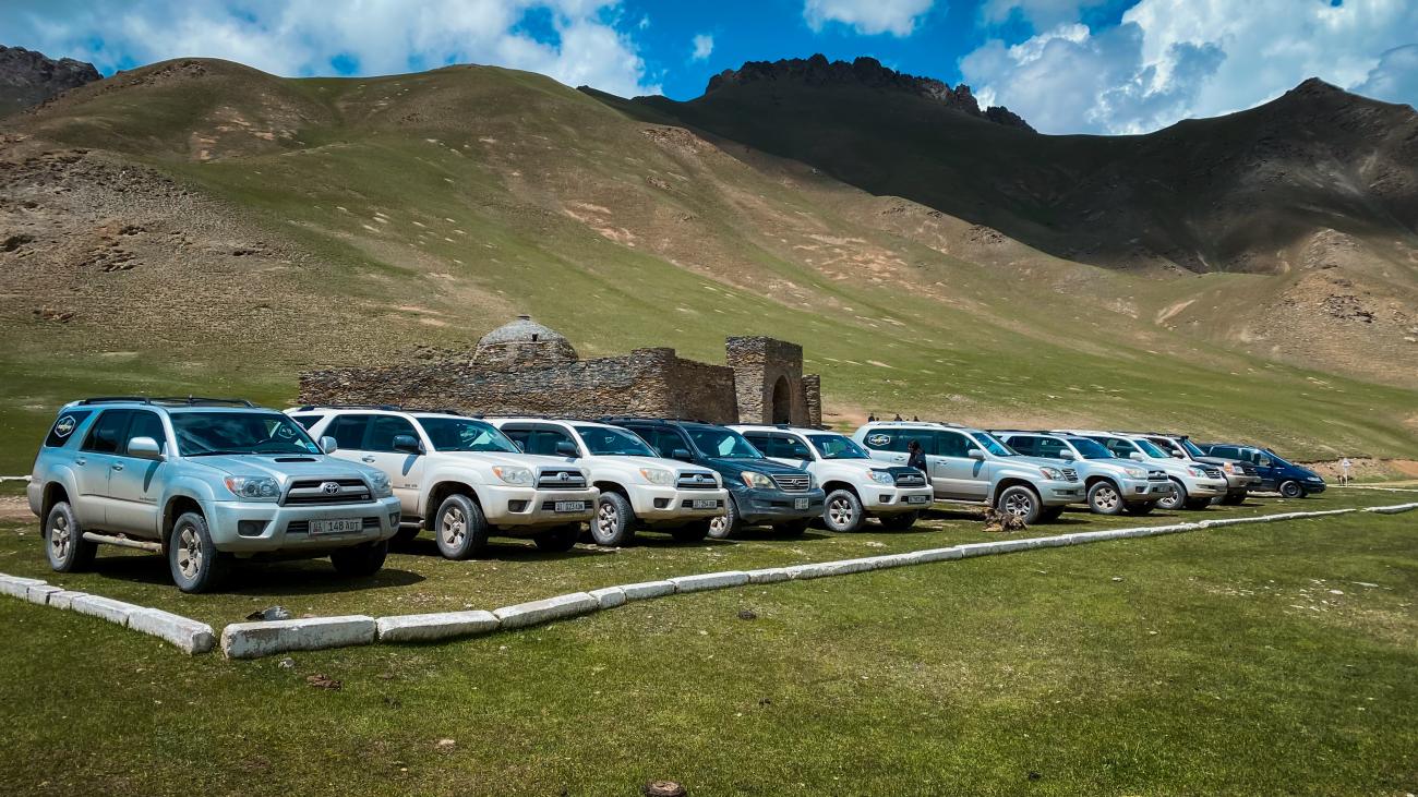 Line of tour SUVs parked in front of the ancient Tash Rabat caravanserai stone ruins in a high mountain valley.