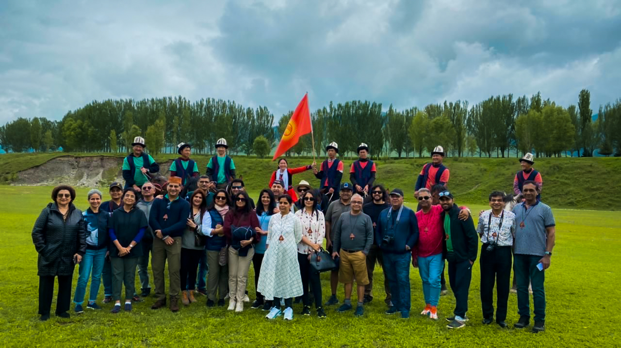 Tour participants and local hosts pose together holding the Kyrgyz flag on a green field lined with poplar trees.