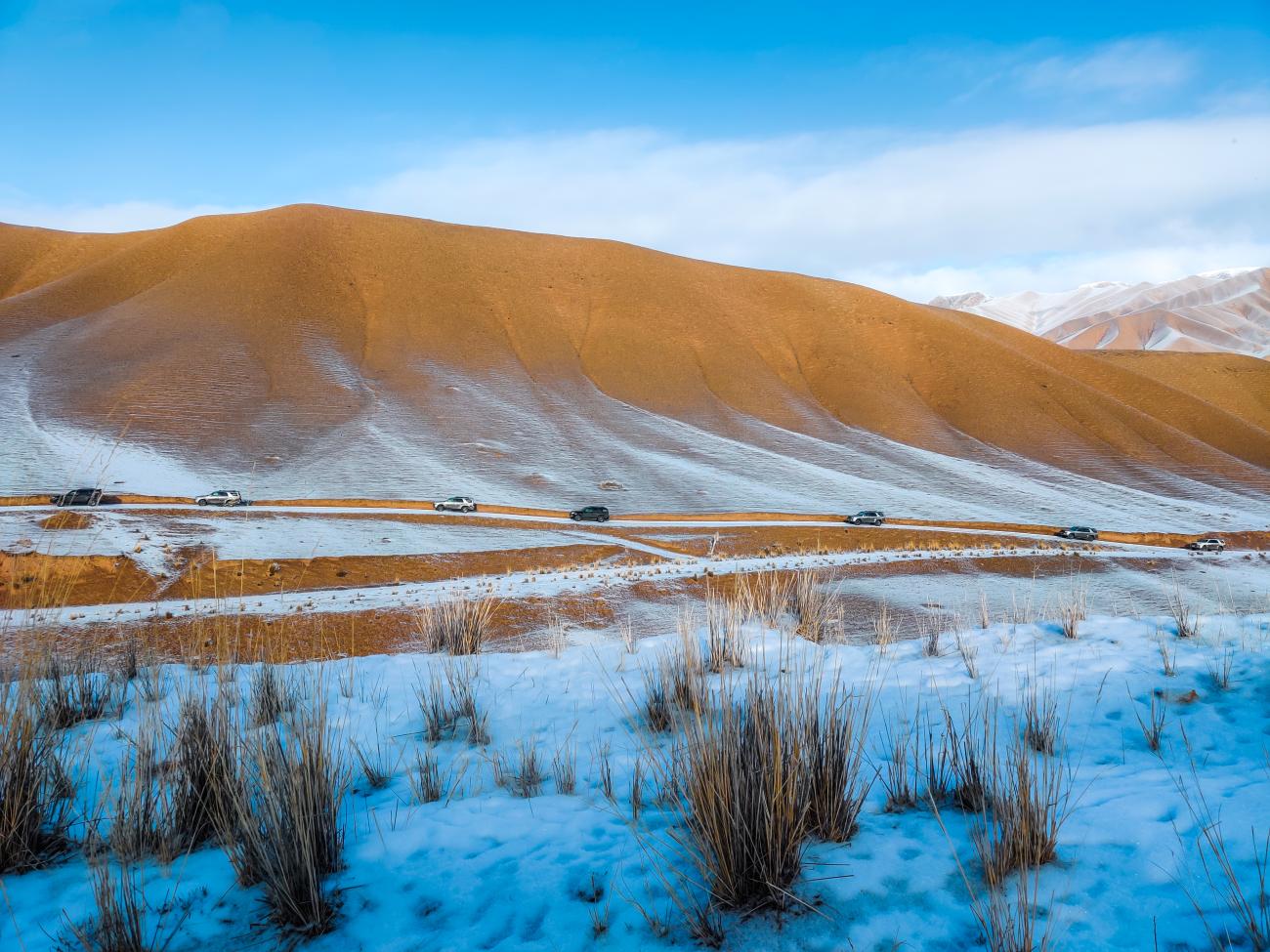 A convoy of SUVs winding along a snowy road beneath golden-brown rolling hills dusted with snow
