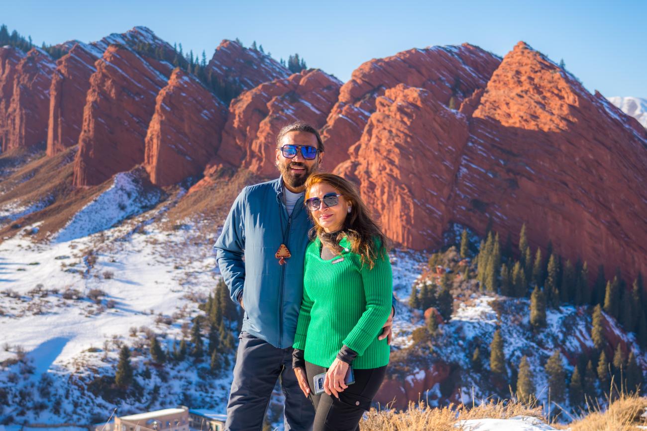 A couple smiling for a photo with the towering red Jeti-Oguz rock formations behind them