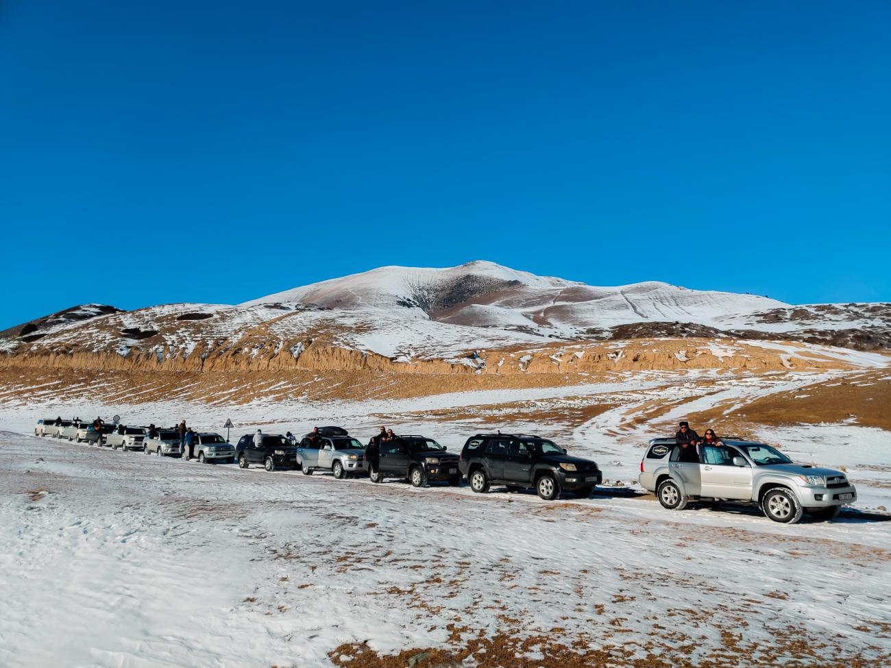 A long line of SUVs parked along a snow-dusted hillside with mountains under a clear blue sky