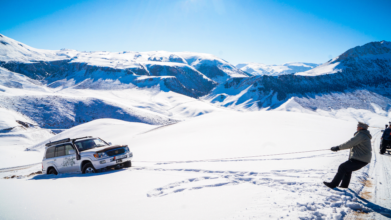 A 4x4 being towed by rope on deep snow with a panoramic backdrop of snow-capped Kyrgyz mountains