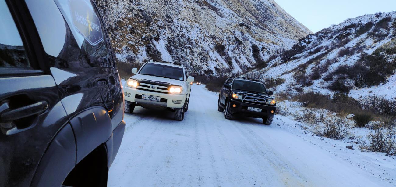 Three SUVs with headlights on navigating a narrow snow-covered mountain pass at dusk