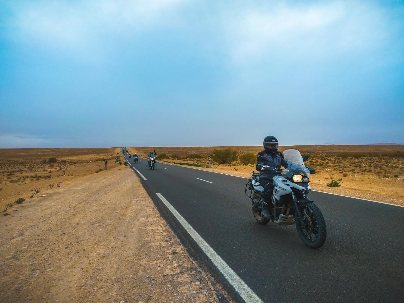 Adventure motorcycles riding in formation along a straight desert highway in Morocco.