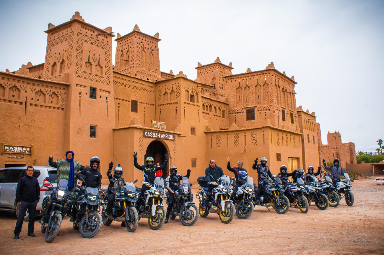 Motorcycle riders waving in front of the ornate entrance of Kasbah Amridil in Morocco.