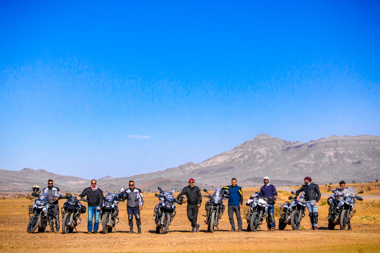A line of riders standing with their motorcycles in the Moroccan desert with Atlas Mountain peaks behind them.