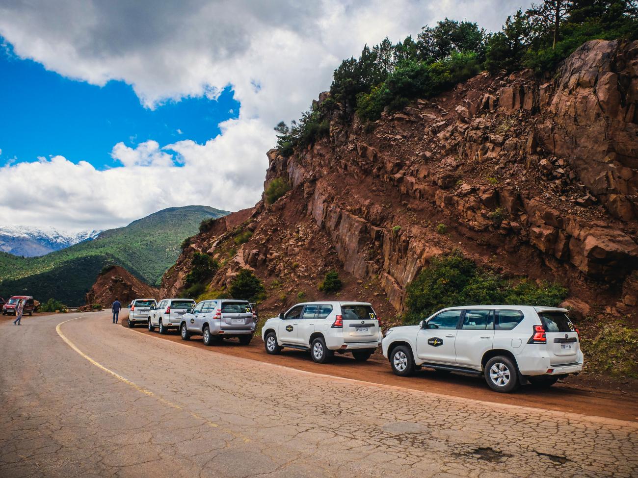 Convoy of white SUVs parked along a winding Atlas Mountains road with red rock cliffs and snow-capped peaks.