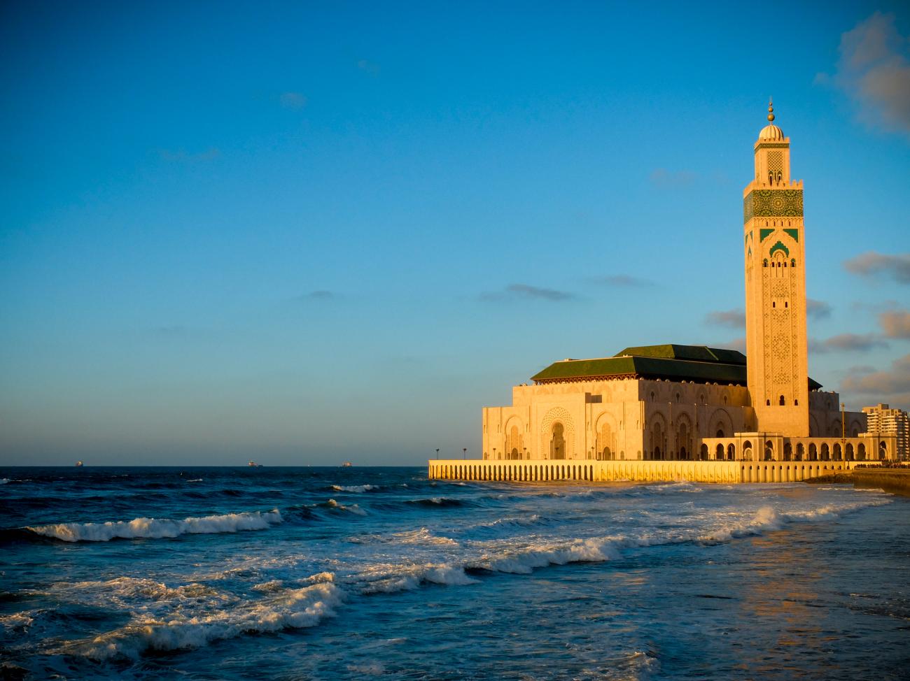 Hassan II Mosque in Casablanca glowing in golden light with Atlantic Ocean waves crashing at its base.
