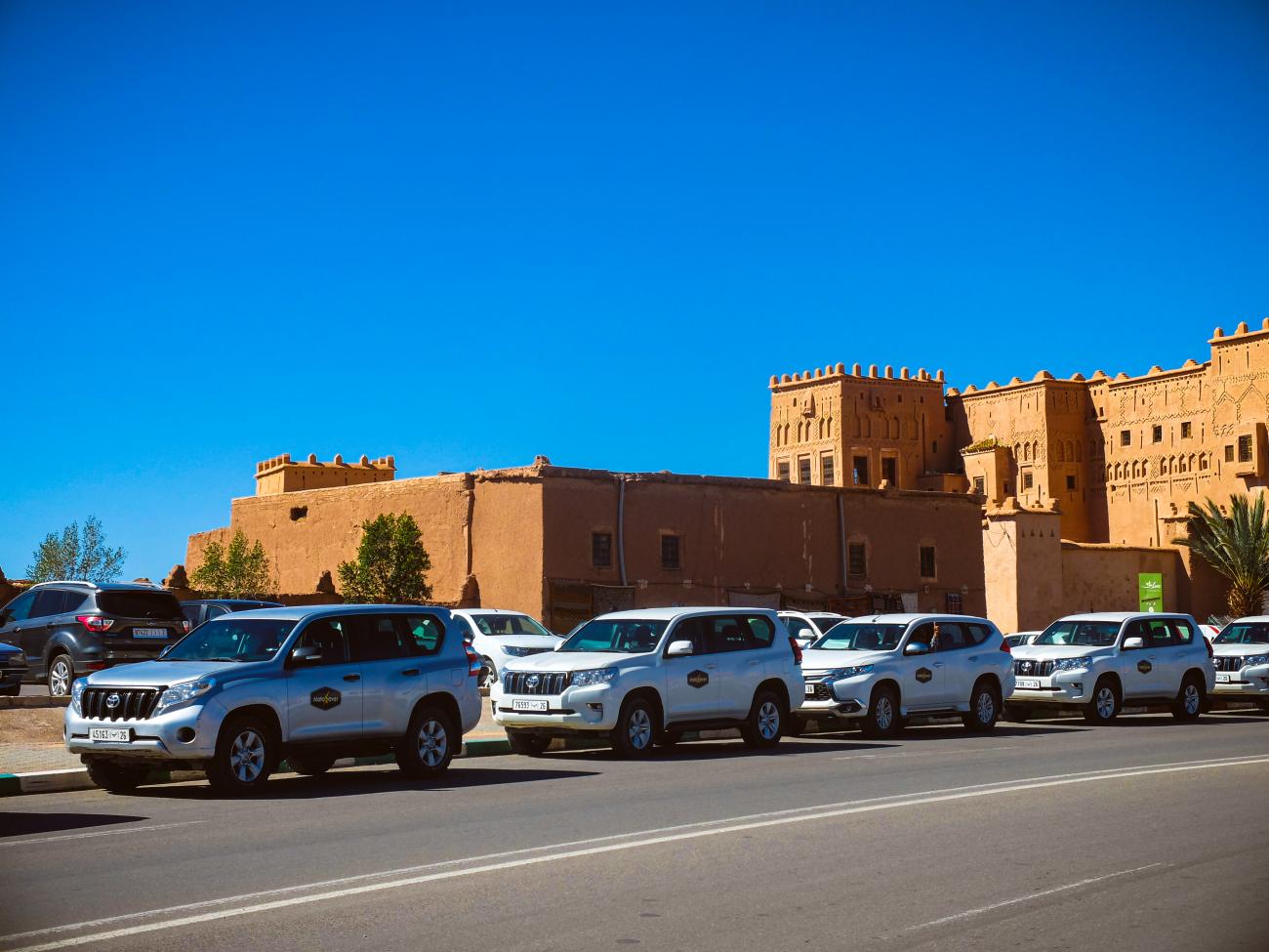 Fleet of tour SUVs parked in front of a historic mud-brick kasbah in Ouarzazate, Morocco.