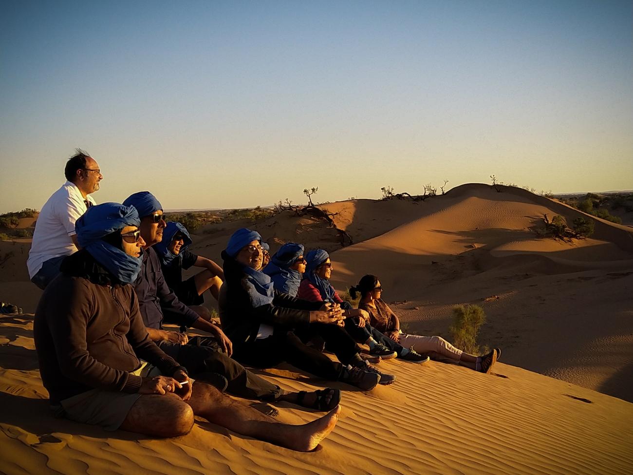 Group of travellers in blue headwraps sitting on Sahara sand dunes watching the sunset.