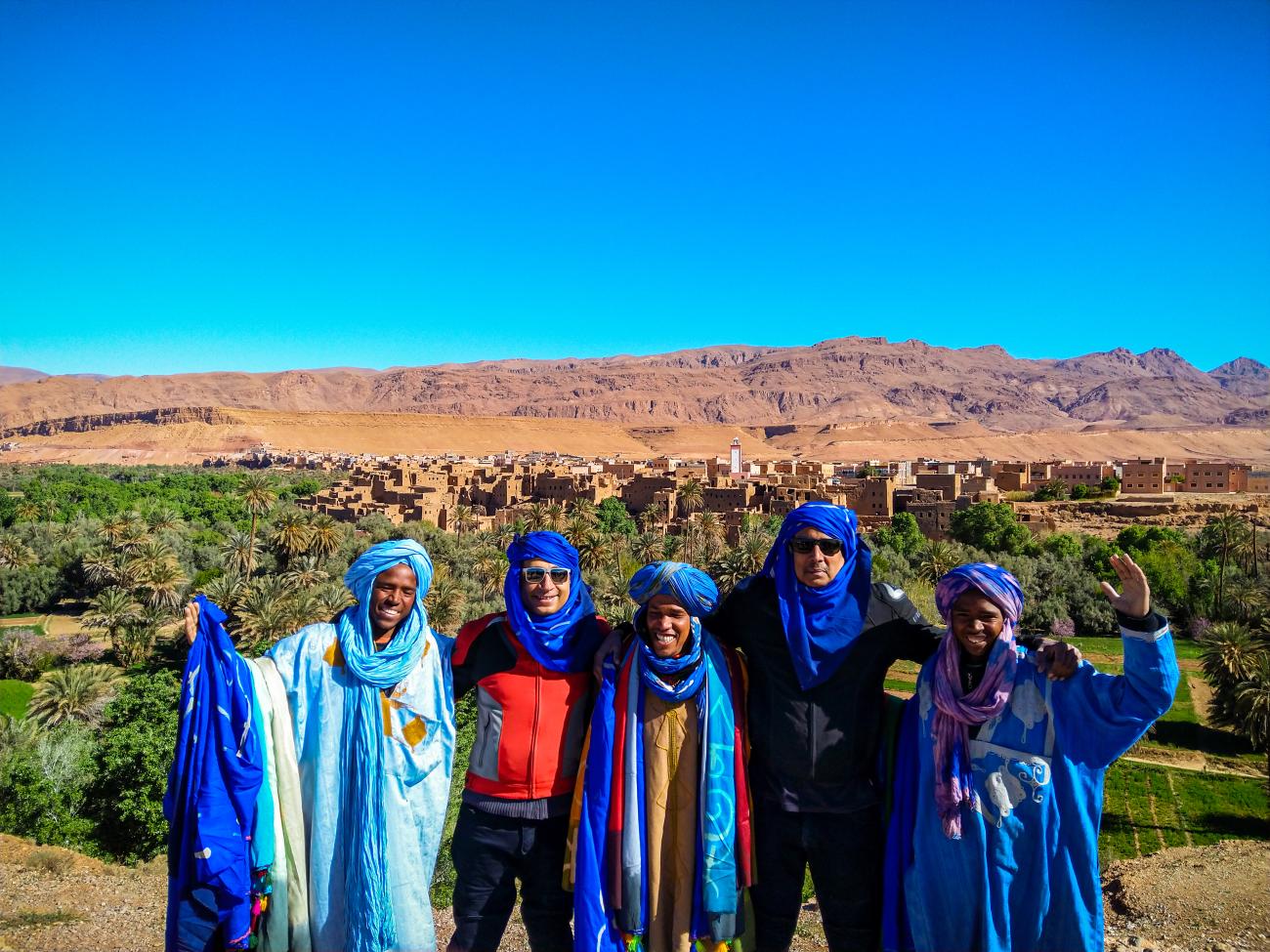 Tour group wearing blue Berber turbans posing with a Moroccan oasis town and mountains in the background.