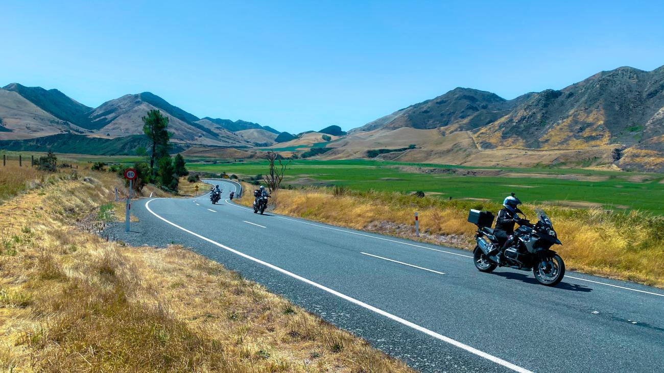 Group of motorcyclists riding through the golden Canterbury hill country with green farmland and brown mountains ahead.