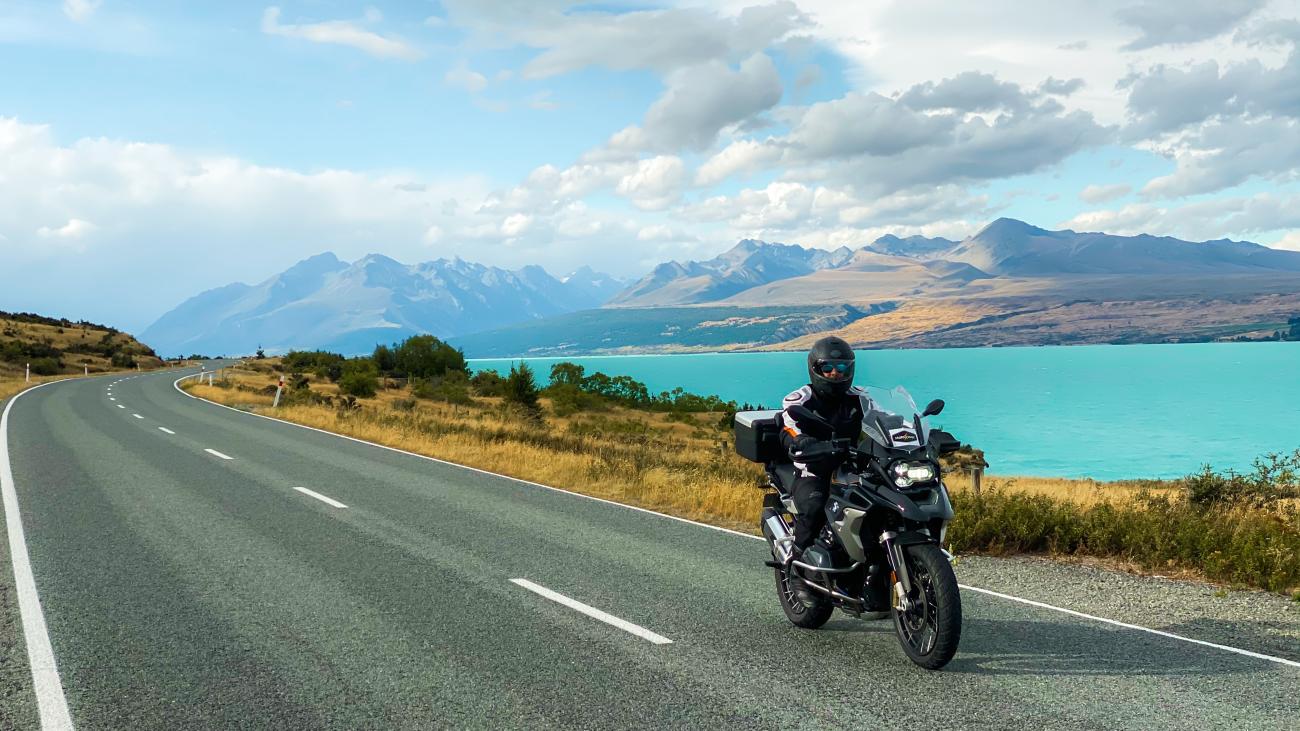 Motorcyclist riding alongside the turquoise Lake Pukaki with dramatic Mount Cook range in the background.