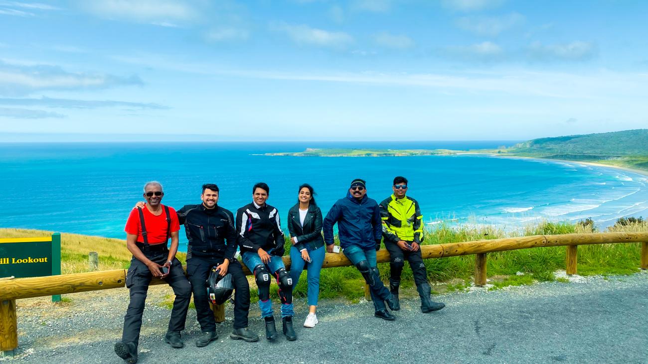 Six MotoRover tour group members posing at a coastal hill lookout with a panoramic view of the turquoise bay below.