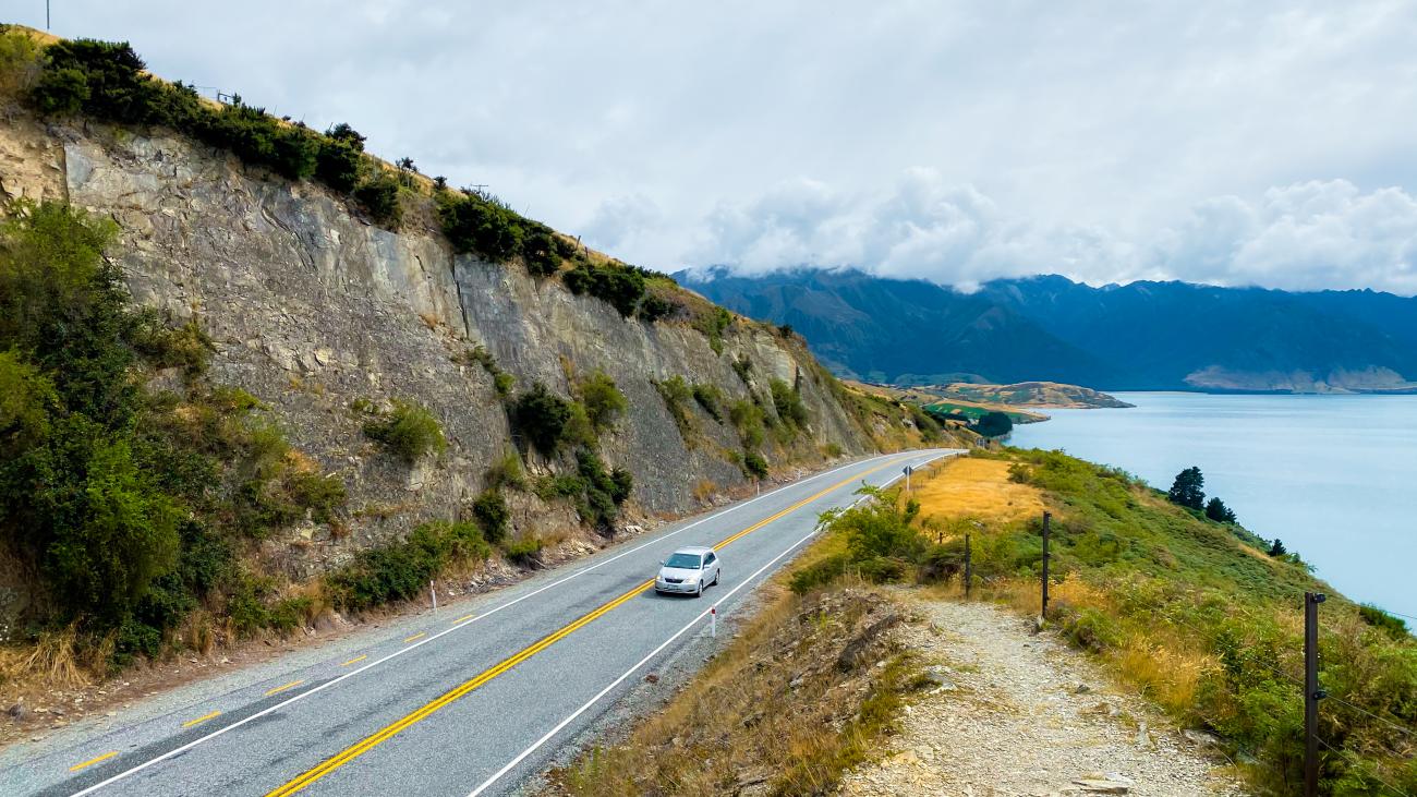 Car driving along a cliff-carved lakeside highway with mountains and overcast skies in New Zealand's South Island.
