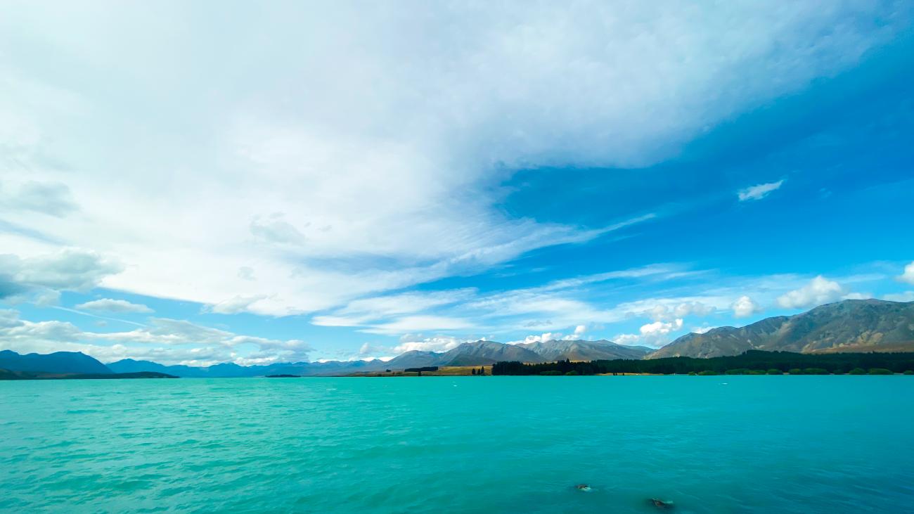 Panoramic view of turquoise Lake Tekapo surrounded by distant mountain ranges under dramatic clouds in New Zealand.