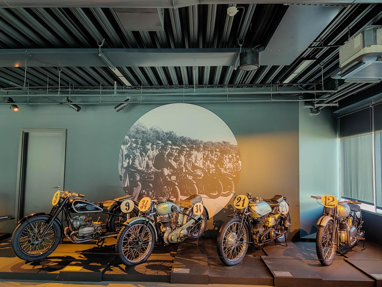 Collection of vintage racing motorcycles displayed against a historic photo backdrop at the Riga Motor Museum.