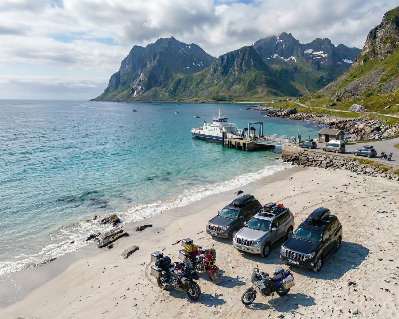 Motorcycles and support vehicles parked on a white-sand Lofoten beach with turquoise water, mountains, and a ferry at the dock.