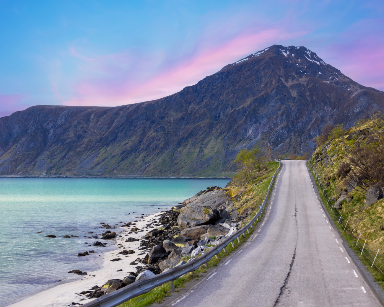 Coastal road stretching along turquoise waters toward a snow-dusted mountain under a pink sunset sky in Lofoten.
