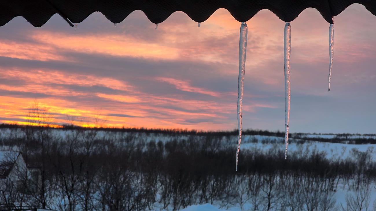 Icicles hanging from a rooftop framing a golden pink sunset over the snow-covered Arctic tundra.