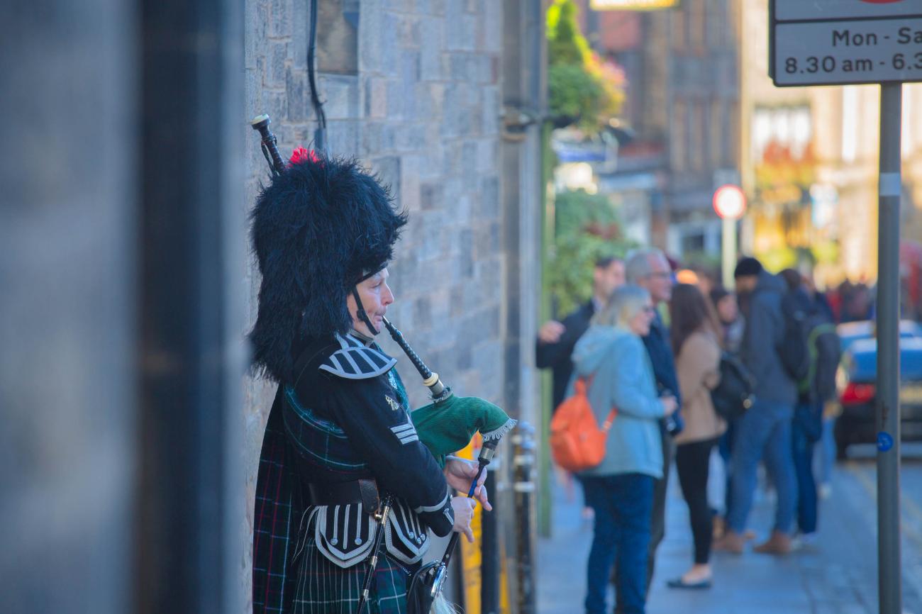 A bagpiper in traditional Highland dress performs on a busy Edinburgh street with pedestrians passing by.