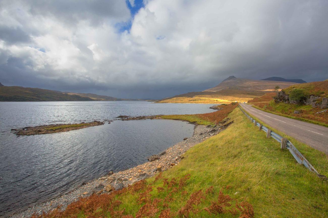 A narrow road runs beside a calm Highland loch with distant mountain peaks under dramatic storm clouds.
