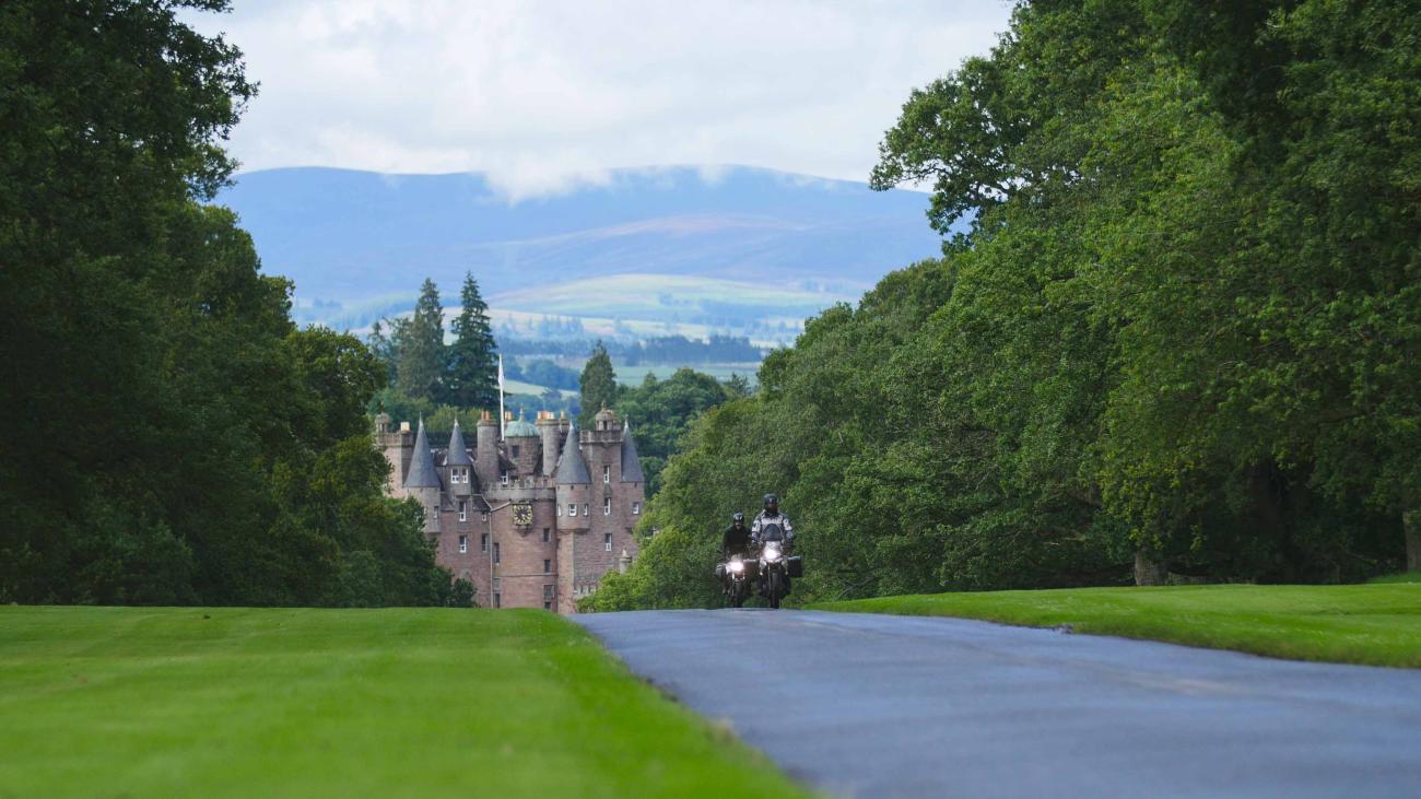 Two motorcyclists ride up a tree-lined driveway toward a turreted Scottish castle with the Cairngorm hills in the background.