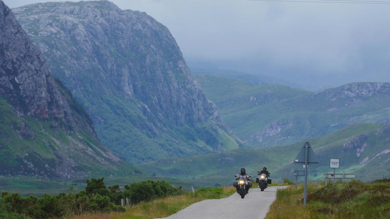 Two motorcyclists approach on a single-track Highland road with a passing-place sign and steep green mountains behind them.
