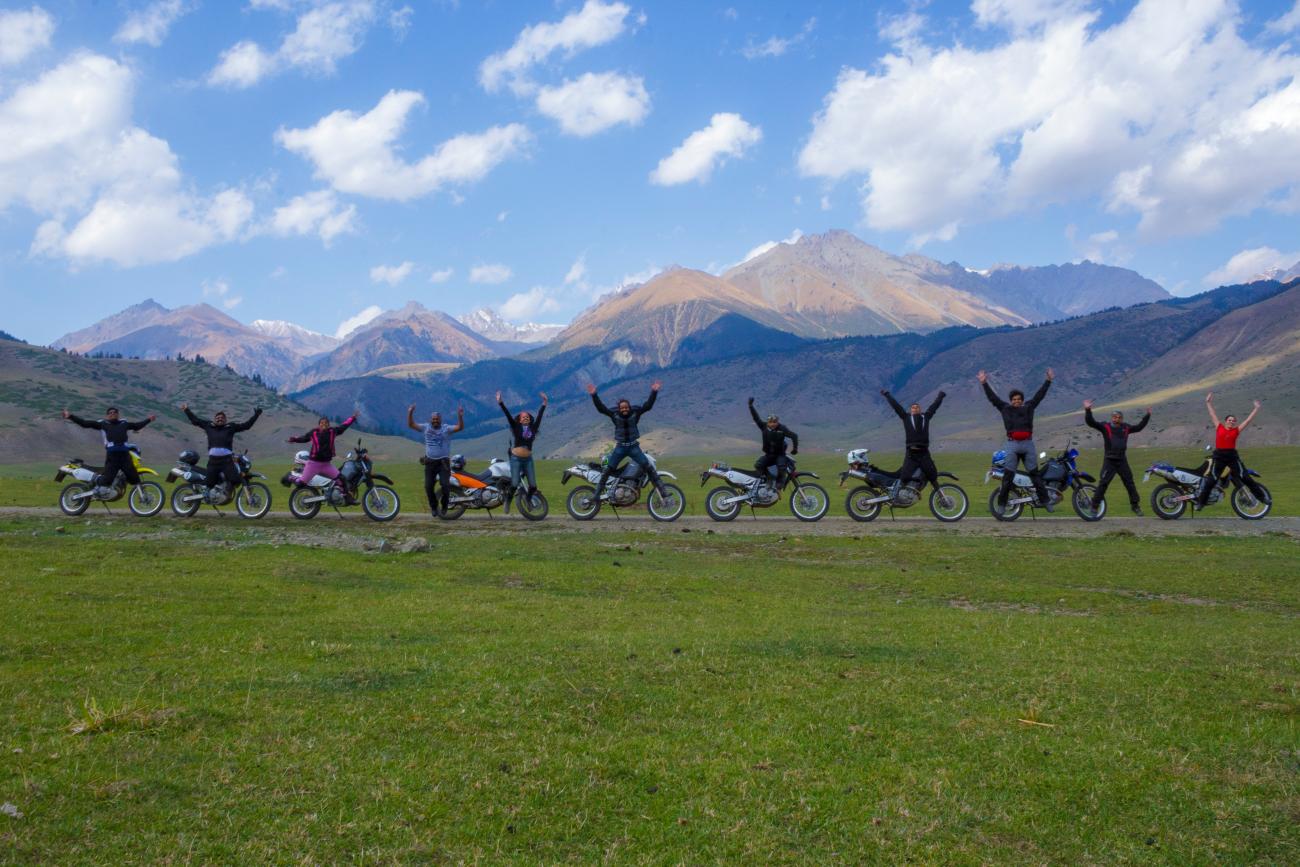 A large group of riders jump and cheer beside their motorcycles on a lush green meadow with snow-capped Kyrgyz mountains behind.