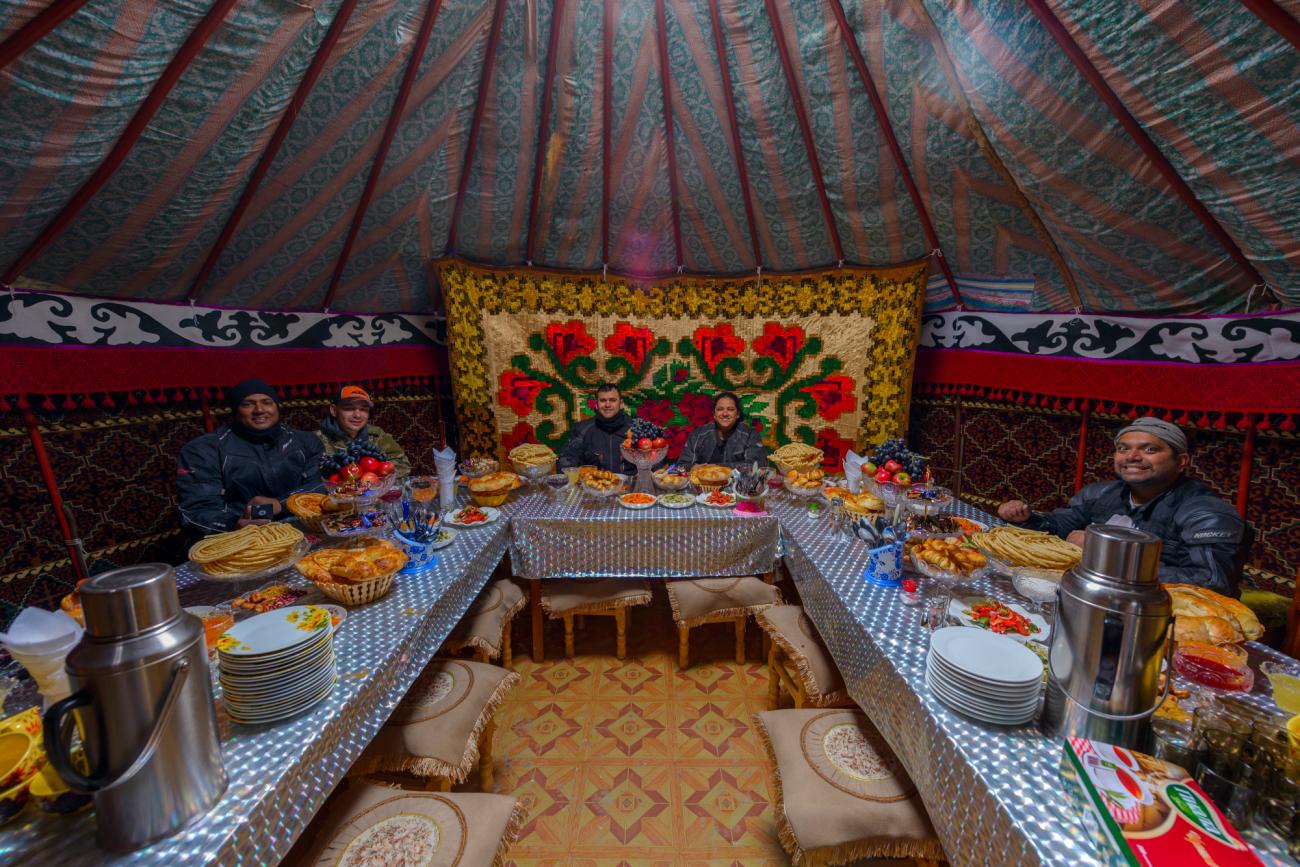 Tour group enjoys a traditional feast inside a colorfully decorated Kyrgyz yurt with bread and local dishes spread on a long table.