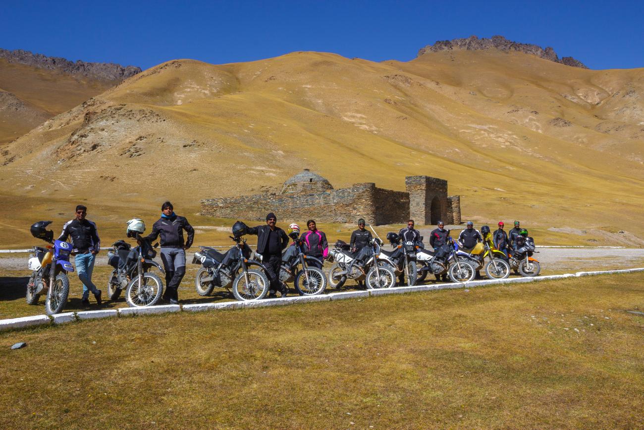 The full tour group lines up with their motorcycles at the Tash Rabat caravanserai under golden hills and blue sky.